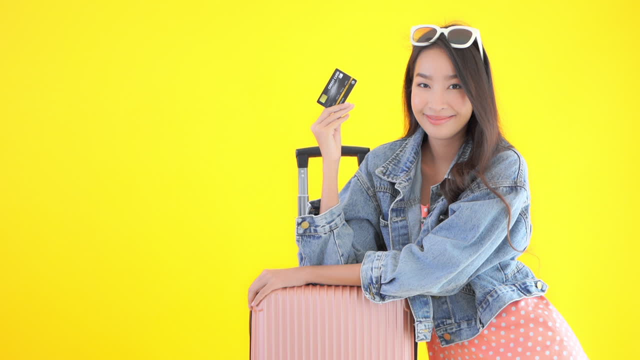 Flight and hotel discount with credit card concept. Asian woman with a pink luggage travel bag showing holding credit card on yellow studio background