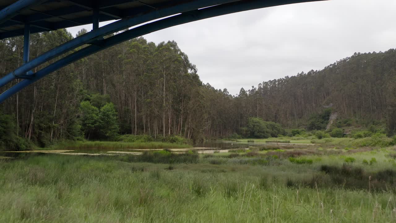 un cautivador drone se desliza bajo el puente de la autopista en san vicente de la barquera, revelando la exuberante vegetación y las tranquilas aguas del río el escudo.