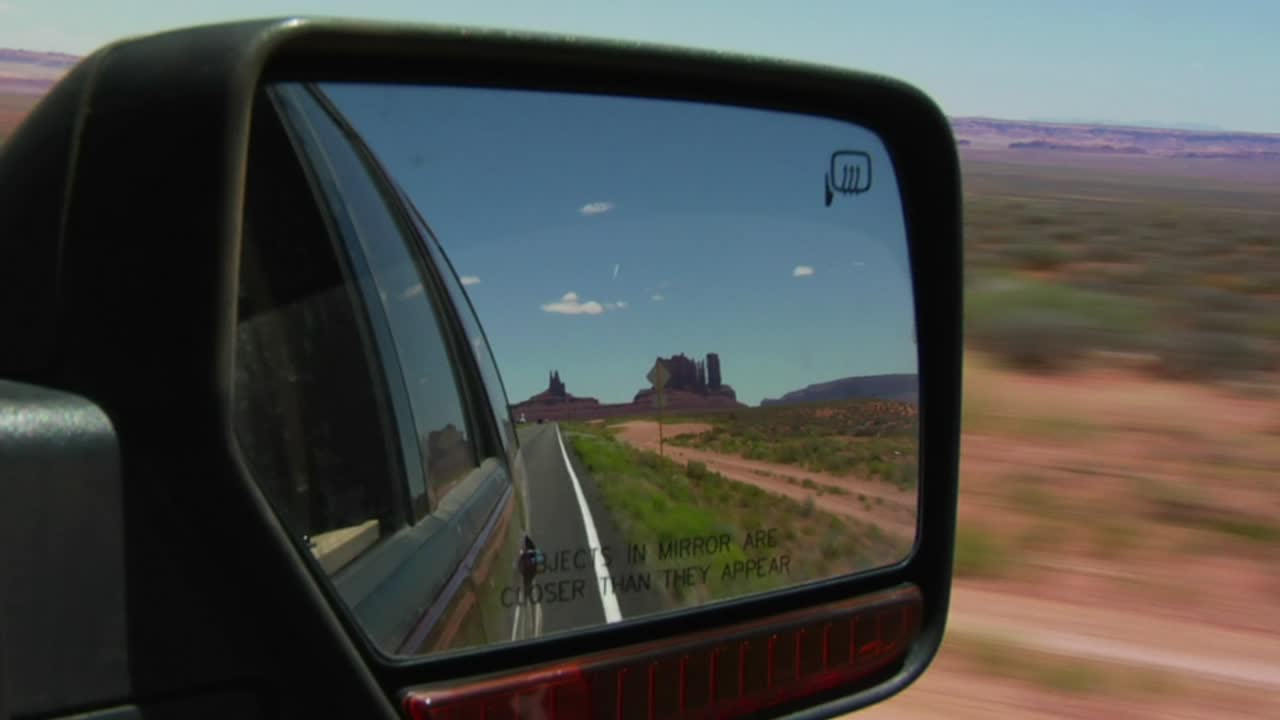 Nahaufnahme eines Außenspiegels eines Fahrzeugs, der eine malerische Wüstenlandschaft mit ikonischen Felsformationen unter klarem blauem Himmel widerspiegelt. Der Spiegel zeigt eine Straße, die zu den Sandsteinfelsen führt