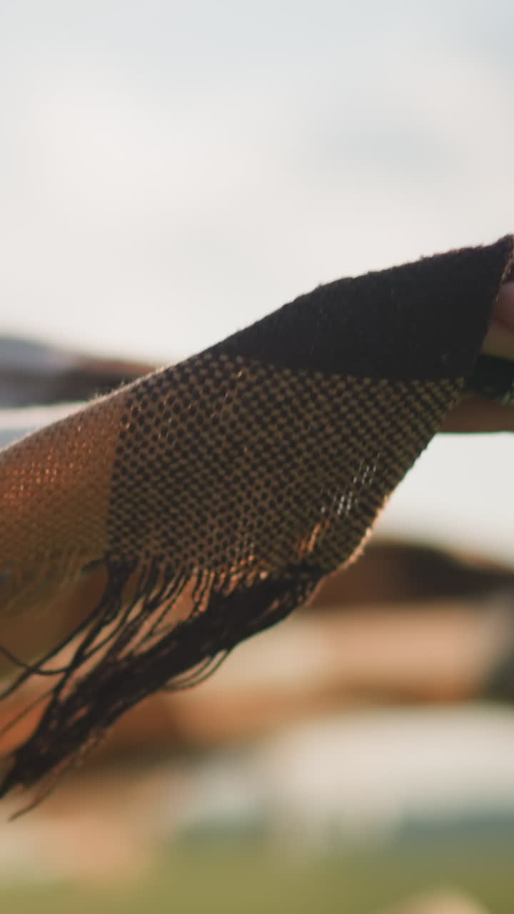 un primer plano de una mano sosteniendo una bufanda a cuadros mientras el viento la sopla suavemente en un campo cubierto de hierba. el fondo está borroso, creando una atmósfera serena y pacífica