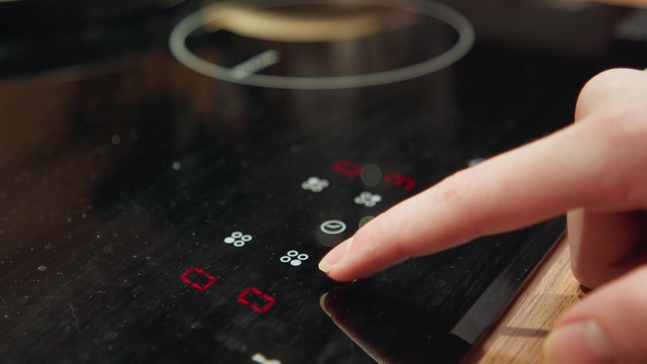 Dito che preme il controllo touch su un piano cottura a induzione nero lucido, con icona di accensione bianca illuminata, indicatori di calore rossi, contorni riflettenti dei fornelli e dettagli eleganti della superficie della cucina.