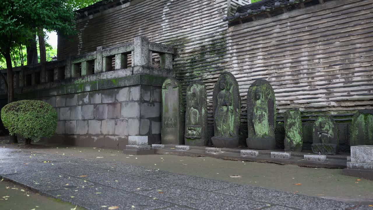 A line of ancient, moss-covered stone statues and monuments stand against a traditional Japanese wall