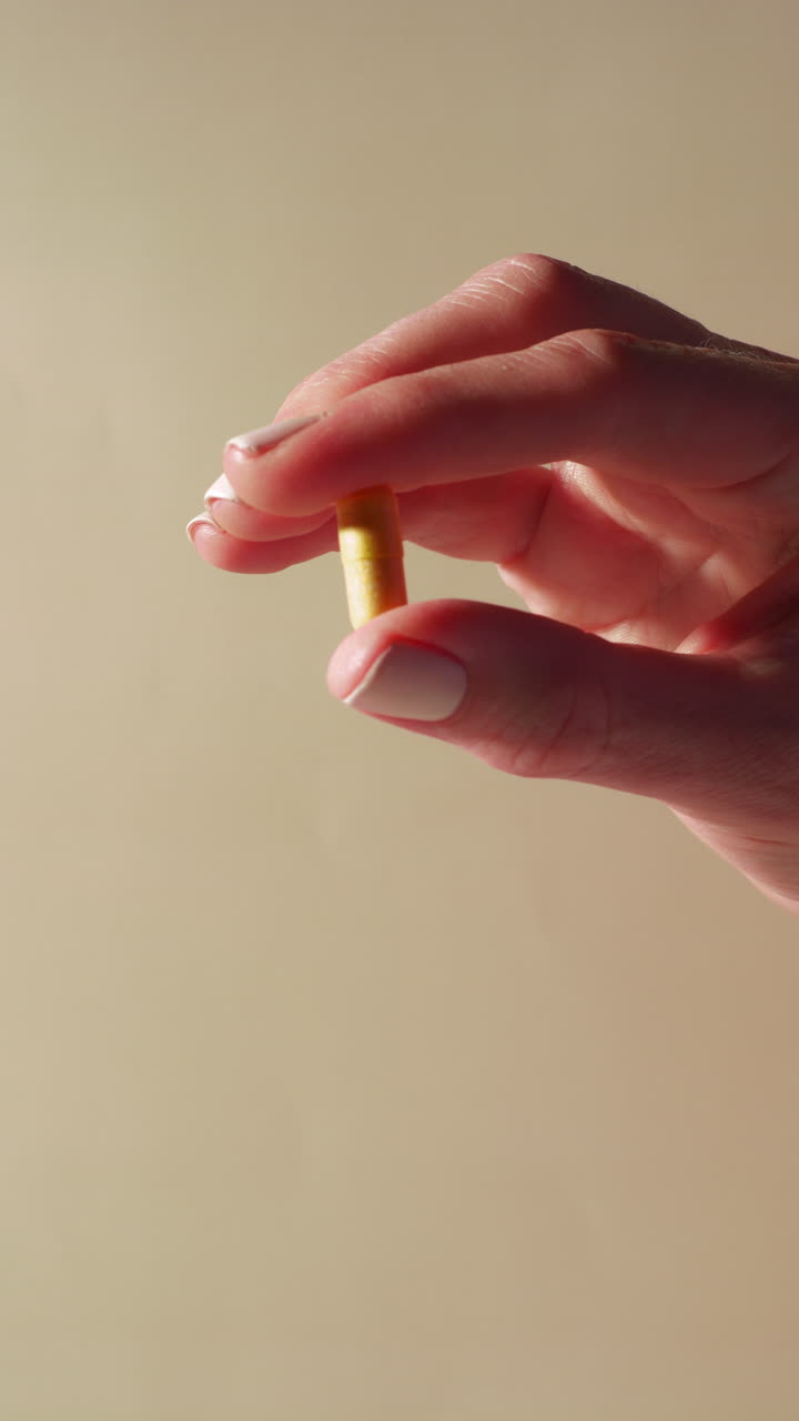 A close-up of a hand gently holding a yellow capsule, possibly a vitamin B complex, cumin, or choline supplement, framed against a neutral beige background with soft daylight highlights.