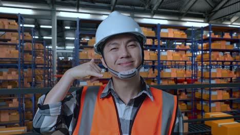 primer plano de un ingeniero masculino asiático con casco de seguridad de pie en el almacén con estantes llenos de mercancías de entrega. sonriendo a la cámara y haciendo un gesto de llamarme en el almacenamiento