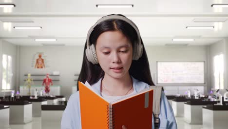 primer plano de una estudiante adolescente asiática con una mochila sosteniendo y leyendo un libro mientras está de pie en un laboratorio de ciencias