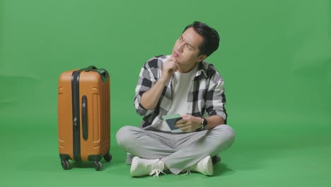 full body of asian male traveler with luggage and passport thinking and looking around then raising her index finger while sitting in the green screen background studio