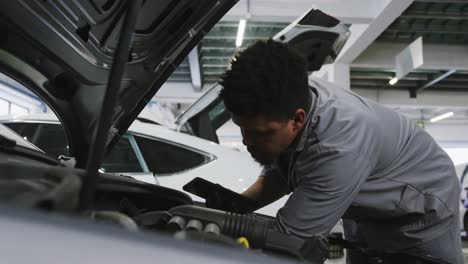 african american male car mechanic looking at an open car engine and using a smartphone