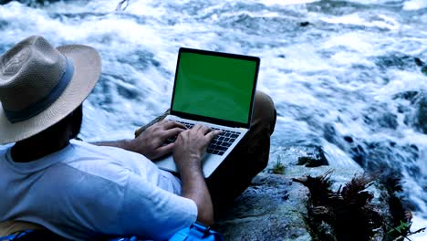 young man using green screen laptop computer on a waterfall. travel and freelance work concept