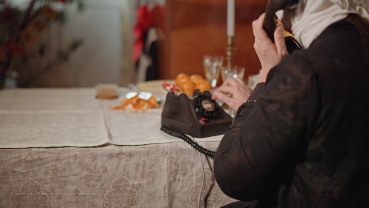 Elderly caucasian woman at table dialing vintage rotary phone, lace tablecloth, tangerines and candlelight, anxious intimate mood as she presses dial and holds receiver, warm domestic interior