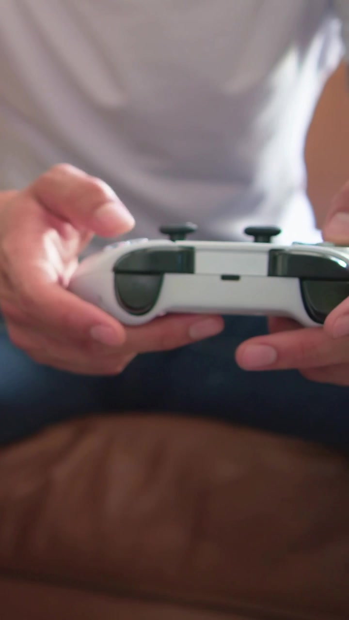 Vertical video: Adjusting grip, man in white tee blue jeans using controller on sofa, seeking win