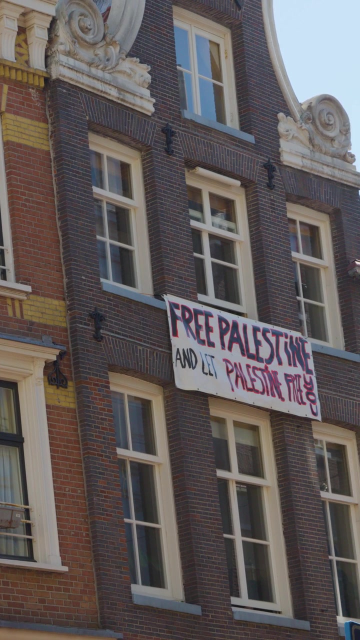 Daytime pan across Amsterdam facades, rainbow flag, and large Free Palestine protest banner visible