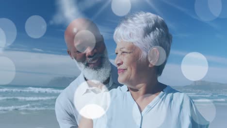 animation of happy senior african american couple embracing at beach over light spots