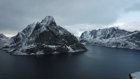 Stunning-drone-flight-over-Reine,-Lofoten-Islands,-Norway,-featuring-snowy-mountains,-the-ocean,-and-a-cloudy-sky