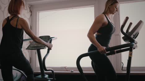 Two-fit-women-exercise-on-cardio-machines-in-gym,-slow-motion