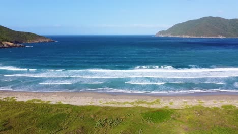 Ein-Lebhafter-Strand-Mit-Rauschenden-Wellen,-Umgeben-Von-Grün-Und-Hügeln,-Unter-Einem-Klaren-Himmel,-Luftaufnahme