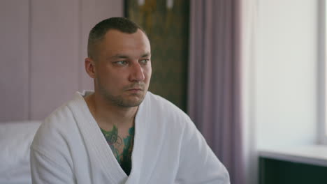 Thoughtful-man-with-tattoos-on-chest-in-bathrobe-sits-on-bed