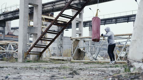 distant view of caucasian man in grey hoodie hitting a punching bag outdoors an abandoned factory on a cloudy morning