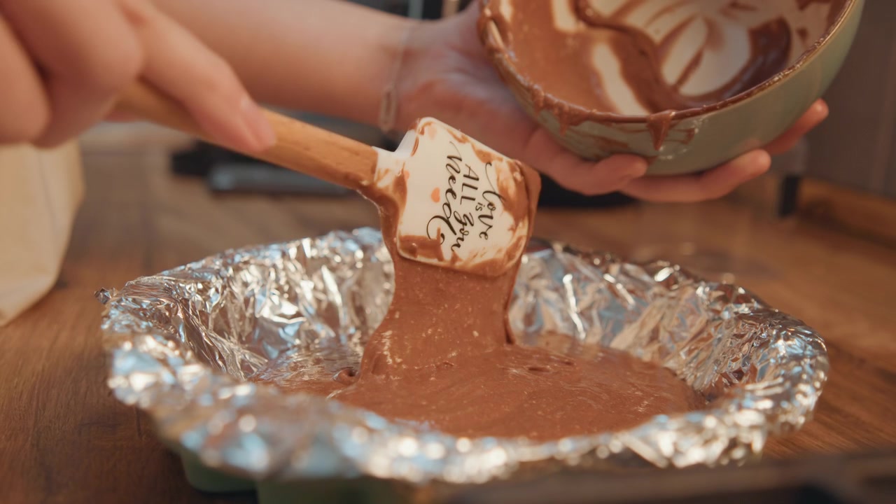 Movimento dettagliato durante la preparazione di dolci. Primo piano di mani che trasferiscono l'impasto al cioccolato in una teglia. Tecnica focalizzata che mostra il movimento mentre l'impasto viene raschiato dalla ciotola in una teglia di alluminio.