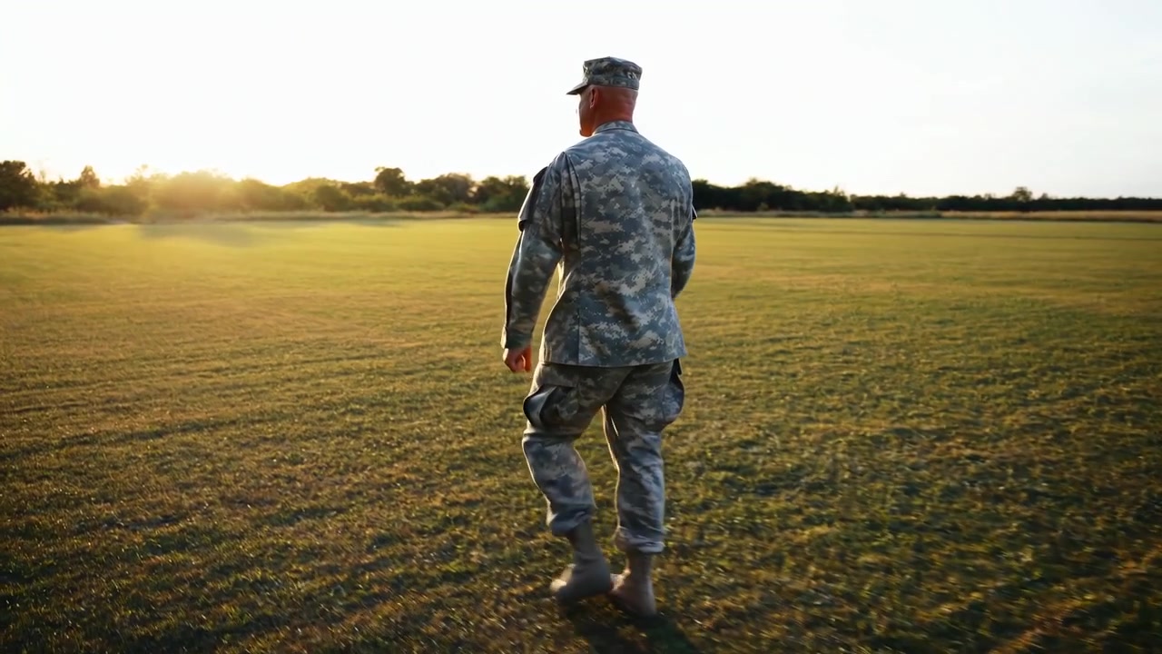 Back view follows a uniformed person striding steadily across an open field while warm low sunlight glows near the horizon, creating long shadows and soft haze around distant trees.