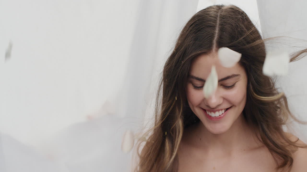 ritratto in slow motion di una bella giovane donna petali di rosa che cadono su una donna attraente che sorride godendo del lusso tra le lenzuola su sfondo bianco concetto di bellezza