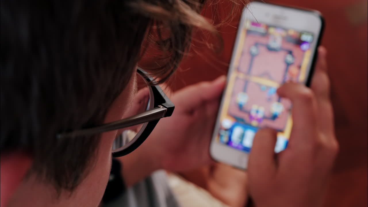 Over-the-shoulder close-up of a kid with glasses playing on a smartphone indoors