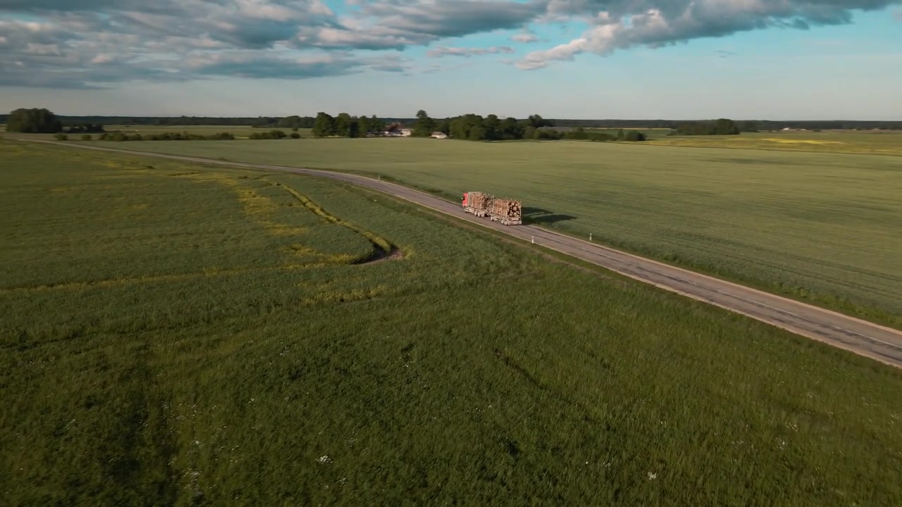 Droneoptagelser viser en tømmerlastbil, der kører til venstre for skærmen på en landevej. Dronen følger lastbilen bagfra og afslører gradvist det vidtstrakte landskab og grønne marker.