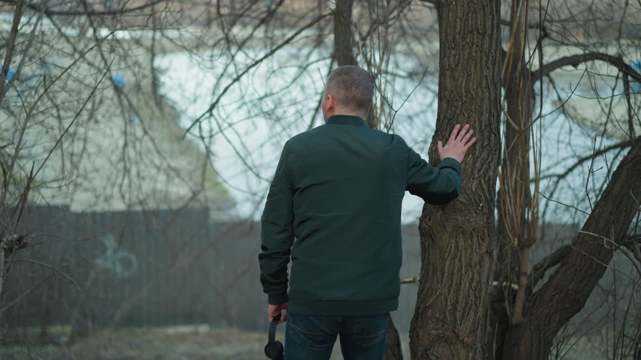 un hombre con una chaqueta verde está de pie en un bosque, tocando la corteza de un árbol con una mano mientras sostiene un par de auriculares en la otra