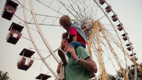 A-little-blond-boy-with-blue-eyes-in-a-red-T-shirt-sits-on-the-shoulders-of-his-dad-with-curly-hair-in-a-Green-T-shirt-in-an-amusement-park-against-the-backdrop-of-a-Ferris-wheel