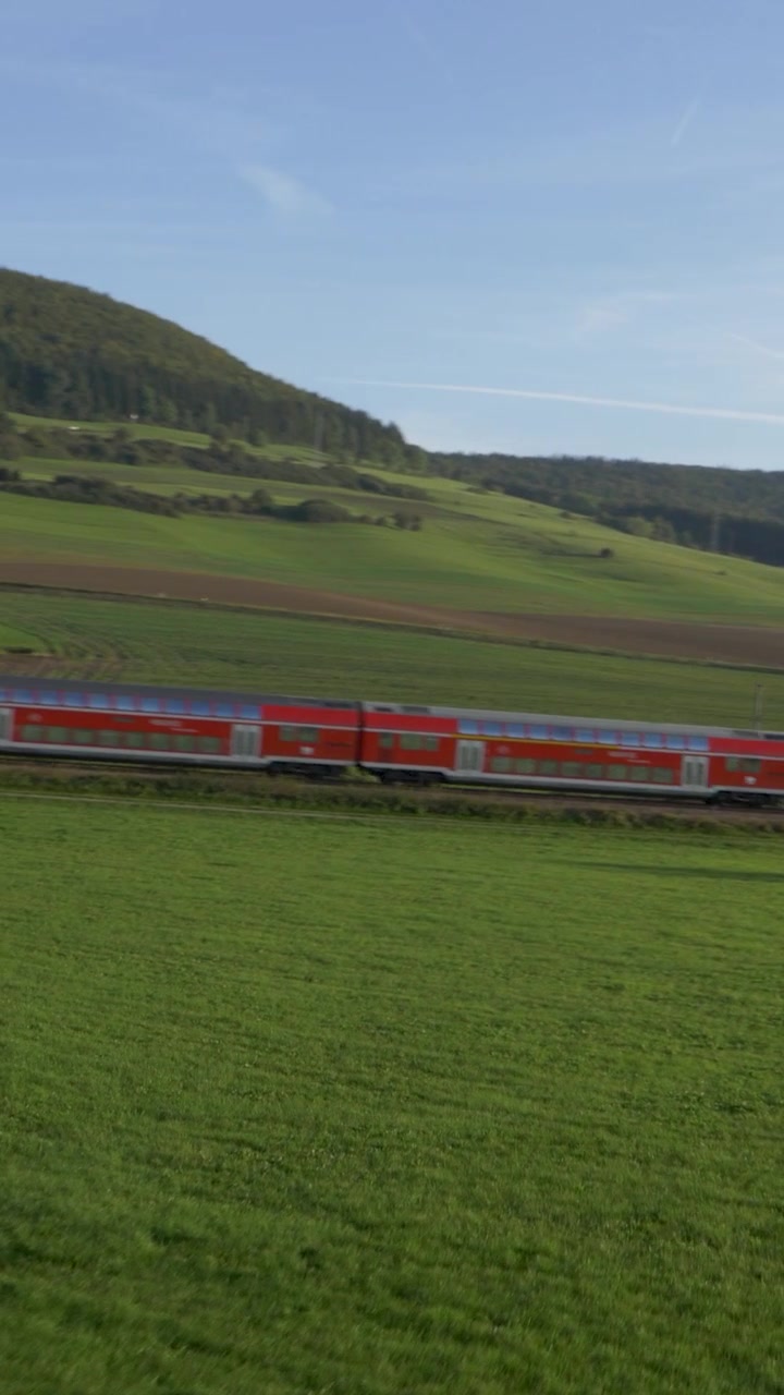 Un treno passeggeri rosso viaggia attraverso verdi campi rurali lungo un fiume sotto un cielo blu.