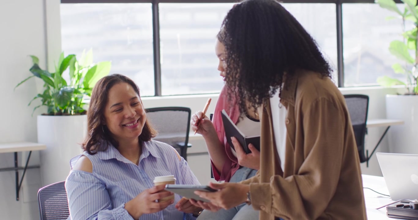 Diverse female coworkers coming to desk by window, showing tablet, notes for review