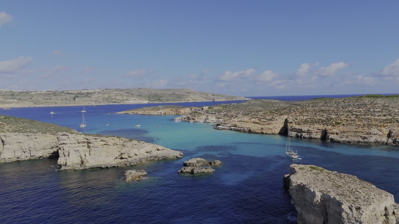 Un fluido volo di drone sopra la Blue Lagoon mostra Comino sulla destra, scogliere rocciose sulla sinistra, acqua turchese calma e diverse barche in una giornata luminosa e soleggiata.
