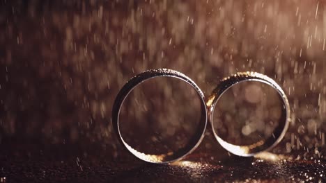 two gold wedding rings under splashes of water on dark surface shining with light close up macro