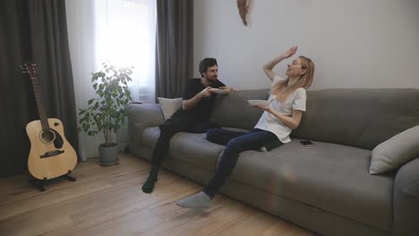 happy couple eating dinner from white bowls, sitting on the sofa and talking