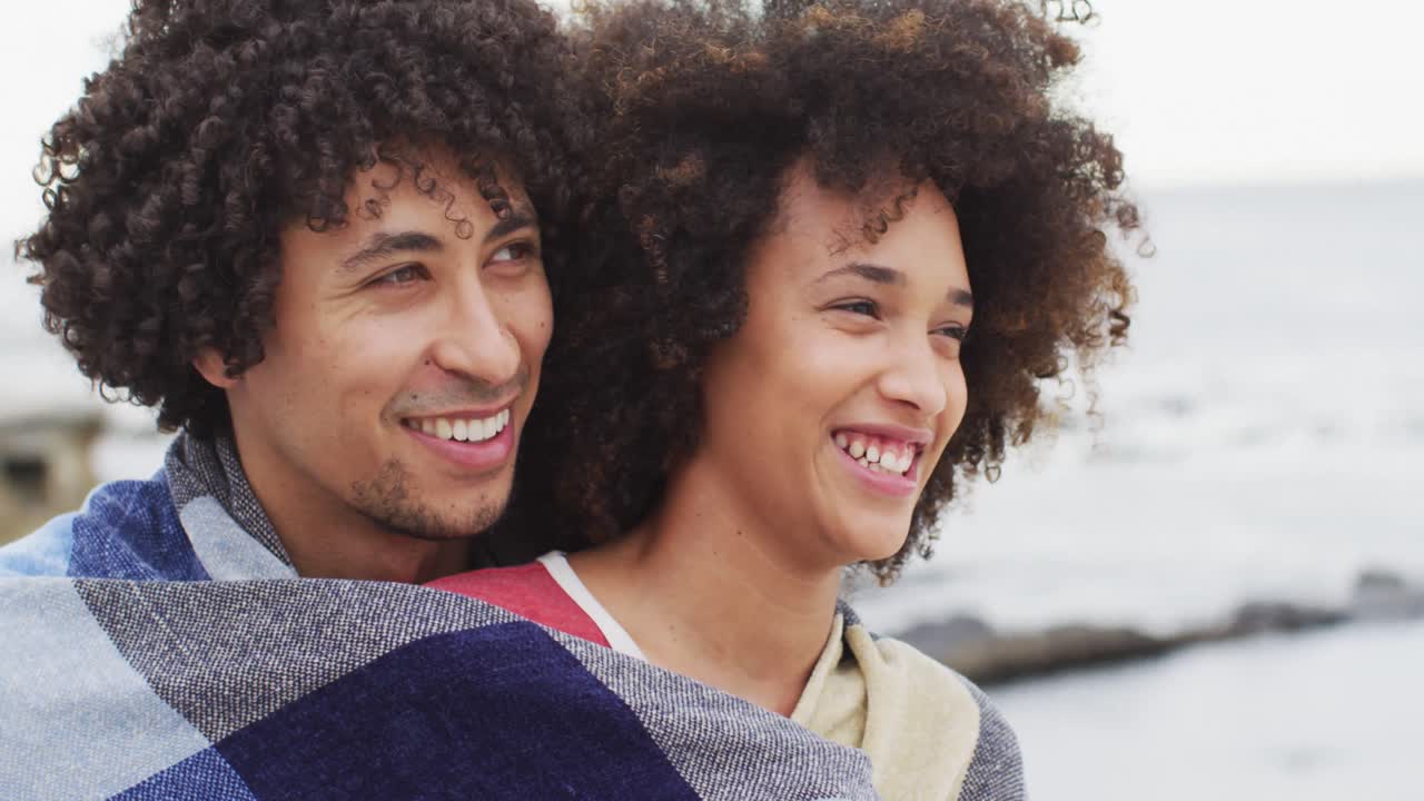 Close up of african american couple wrapped in blanket smiling on the promenade near the beach