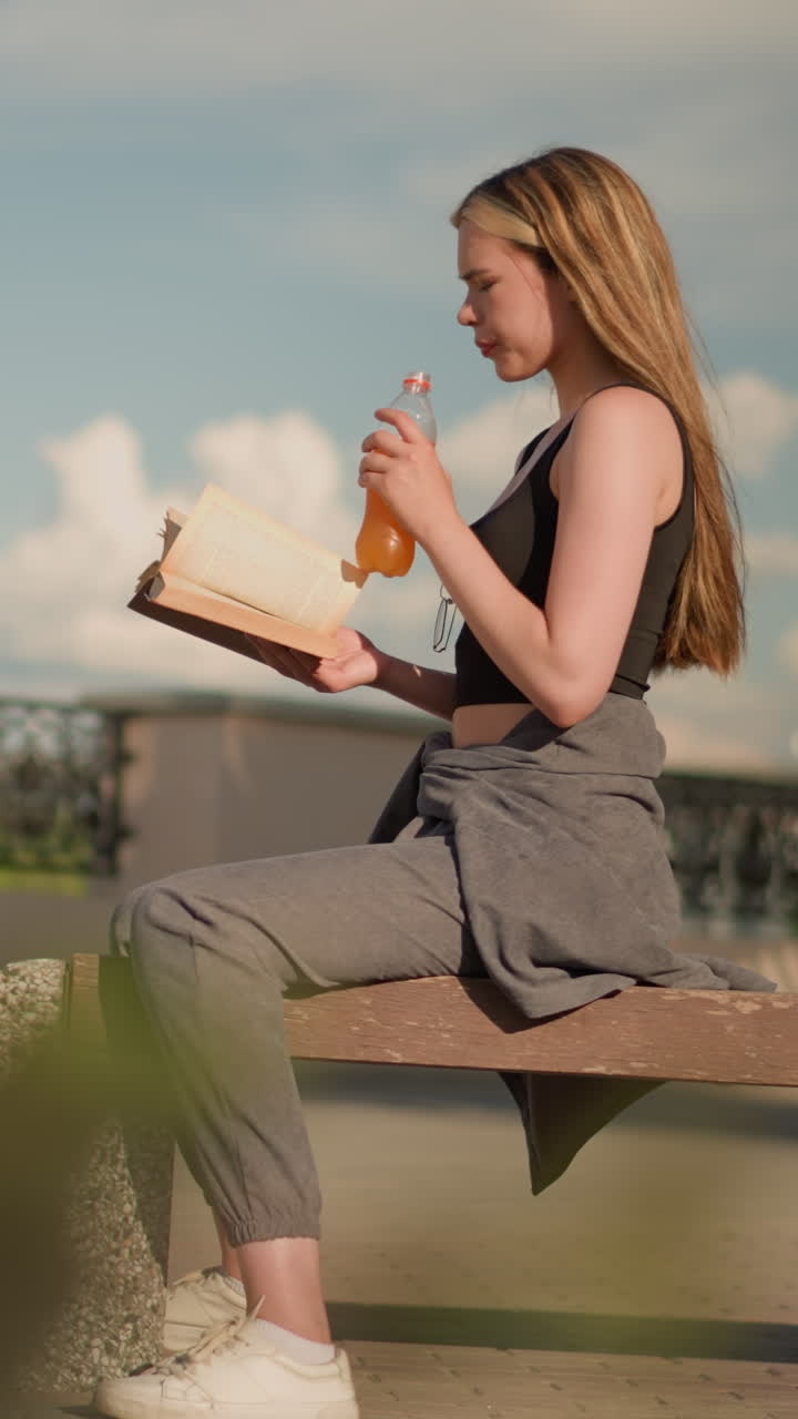 Woman seated outdoors, holding book in one hand while sipping from juice bottle with the other, she continues reading while casting shadow on floor in warm daylight, with background fence