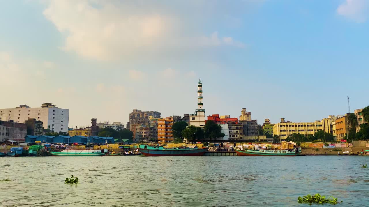 Dhaka city view from Buriganga river in Bangladesh. A dragonfly flying over the river water. Several boats are visible, along with a mosque minaret and city buildings.