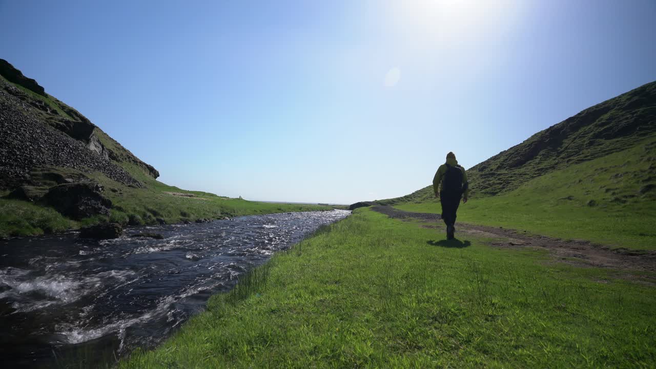 un hombre caminando por un camino de tierra junto a un pequeño arroyo en un hermoso valle verde de montaña en un día soleado