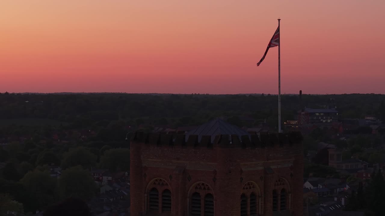 Aerial drone orbit captures the England flag waving atop St Albans Cathedral during sunset, with warm orange light and the city skyline creating a majestic and patriotic scene
