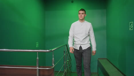 front shot of dude in shirt and trousers walking up stairs, moving into billiard room. confident stride with serious expression. green lighting casts an ambient glow. hand touches green pool table