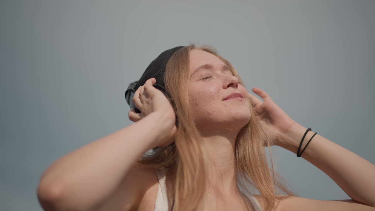 Giovane donna che ascolta la musica con le cuffie sotto un cielo azzurro aperto in una strada tranquilla, occhi chiusi e sorriso, leggero dondolio e postura rilassata, capelli illuminati dal sole e abbigliamento estivo casual, mentre si gode il ritmo e la libertà