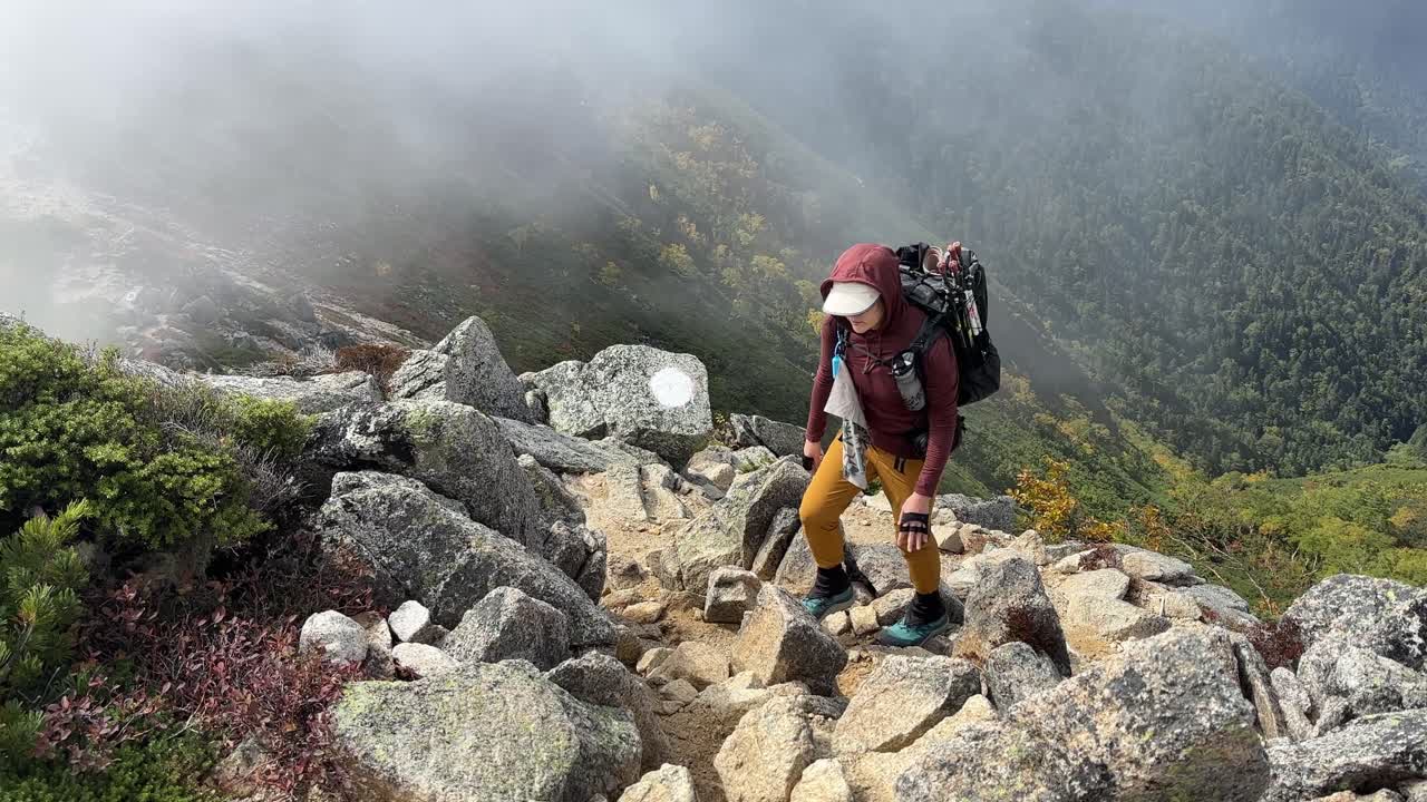 Mujer senderista escalando las empinadas y rocosas laderas del Monte Jonen en Nagano, Japón. La niebla envuelve el escarpado paisaje, revelando el follaje otoñal en el Parque Nacional Chubusangaku.