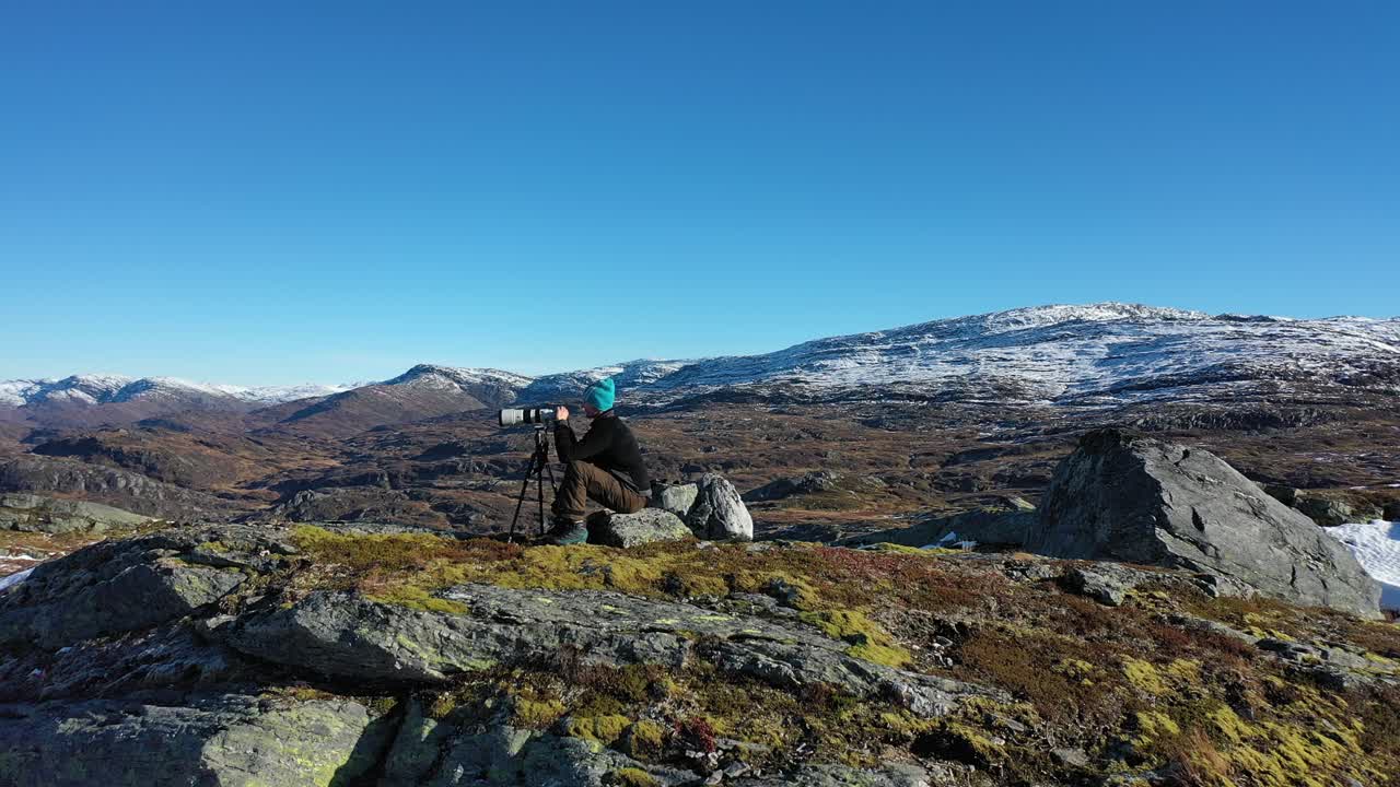 시차 효과가 있는 멋진 가을 산 풍경에서 풍경 사진가 주변의 아름다운 궤도 - 눈 덮인 봉우리와 삼각대에 텔레줌 카메라를 사용하여 바위에 앉아 있는 남성 사진사