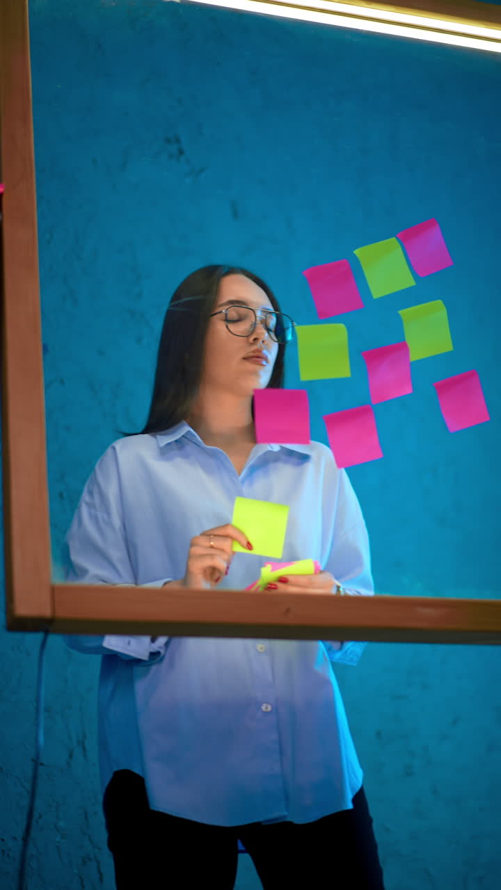 Confident concentrated young Caucasian female takes stickers off the glass board. Ideas concept, business strategy. Vertical video.
