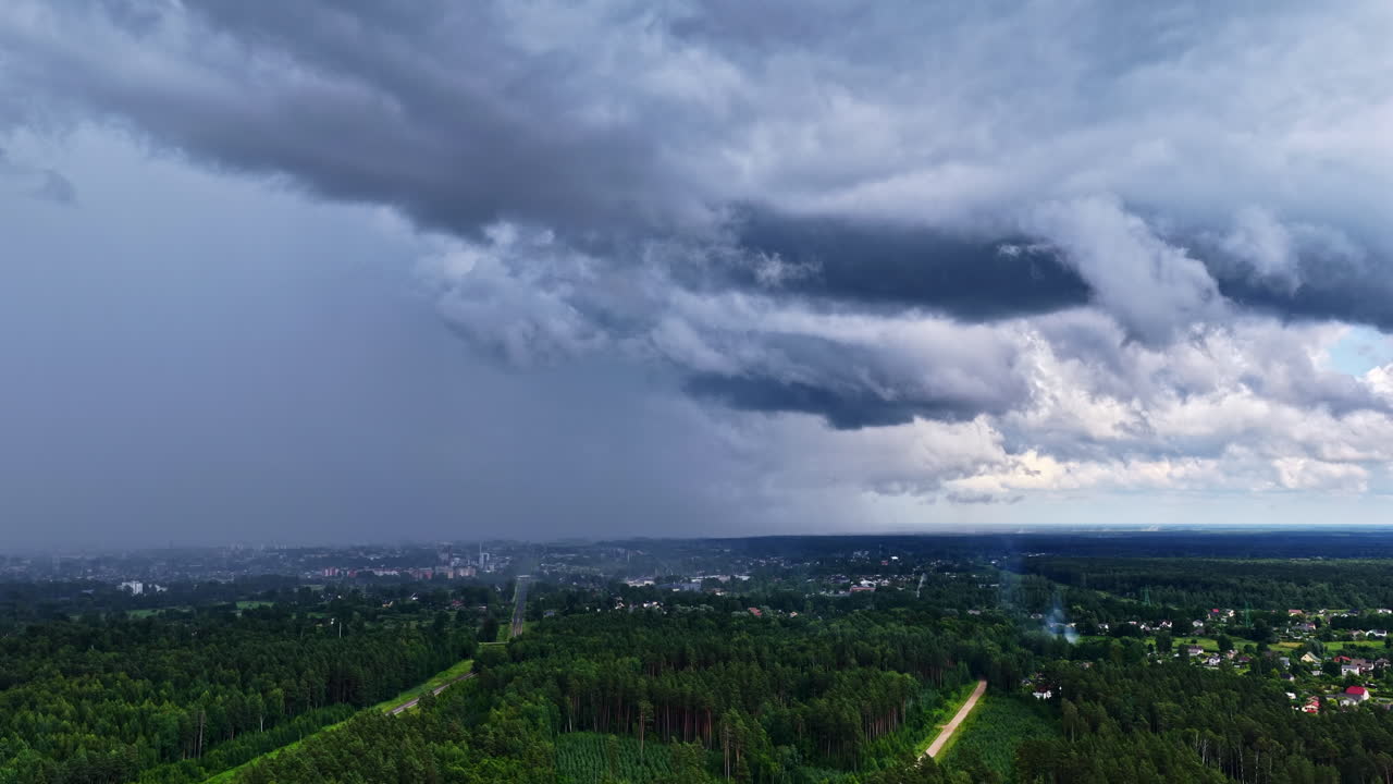 A slow aerial drone shot descends over a dense pine forest, revealing the town of Engure, Latvia, under a dramatic and moody sky filled with dark storm clouds