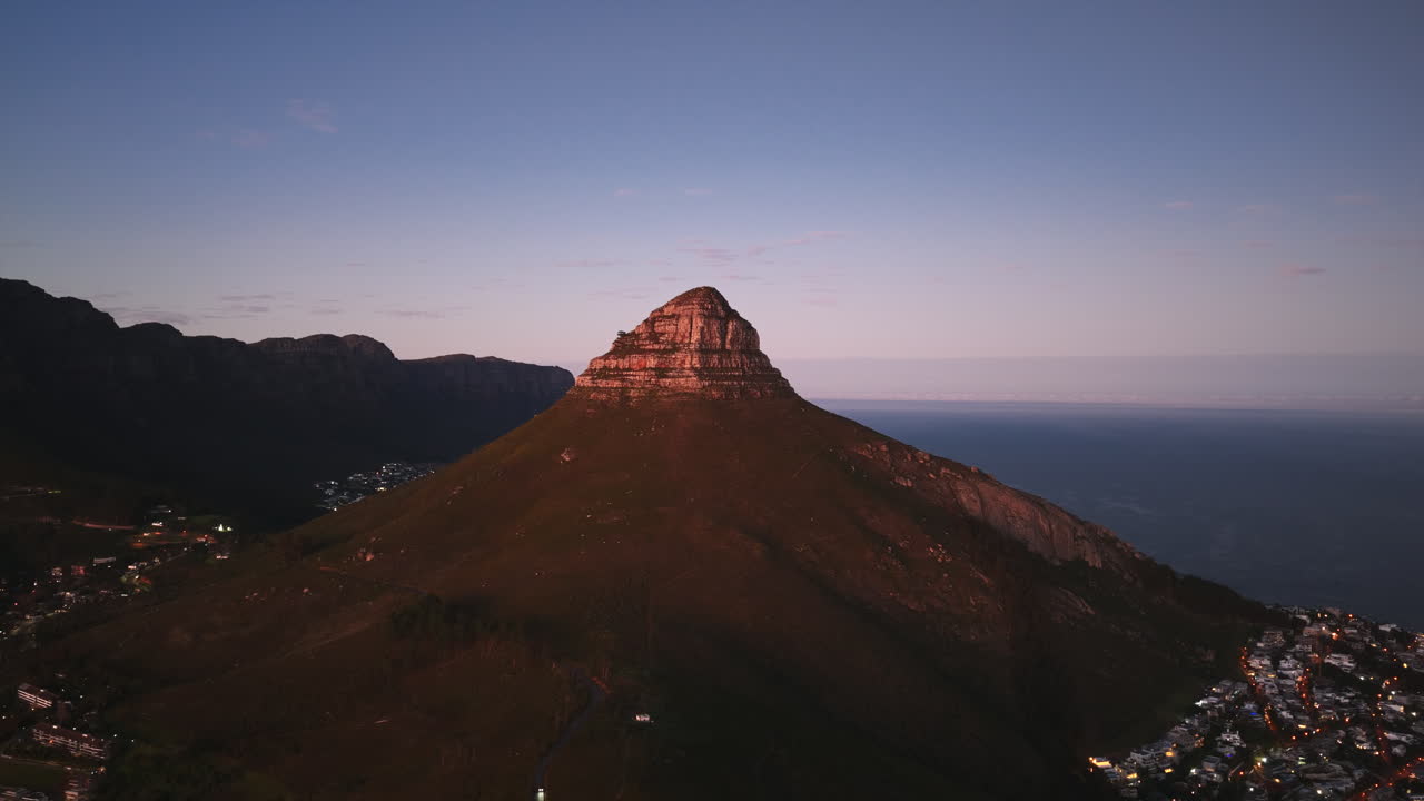 A wide drone view reveals Lion’s Head in its entirety, framed by ocean waters and neighboring peaks, with Cape Town’s suburbs at its base