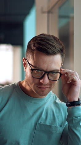 confident good-looking man leaning on the window frame indoors. mid-aged smiling caucasian man puts on glasses and looks at camera. close up. vertical video