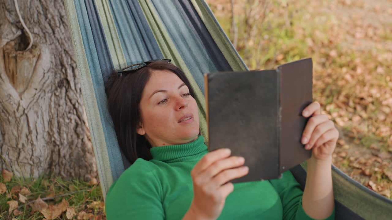 Mulher caucasiana lendo na rede no outono, sob uma árvore, usando suéter verde e óculos de sol, folhas espalhadas pelo chão, virando as páginas de um livro, expressão relaxada, luz solar quente e sombra suave, aconchegante.