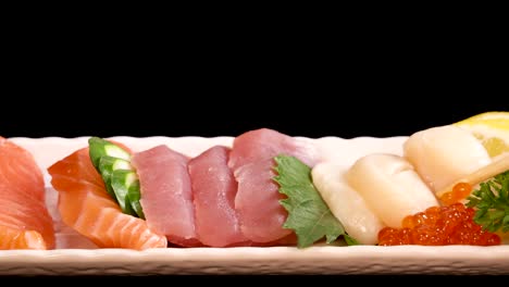 hand uses chopsticks to pick salmon and scallop sashimi from a neatly arranged sushi platter, under bright studio lighting with a black background