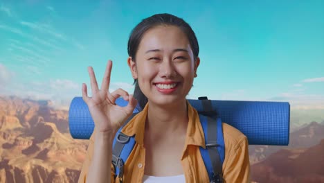 close up of asian female hiker with mountaineering backpack smiling and showing okay gesture to camera while traveling at the top of mountain