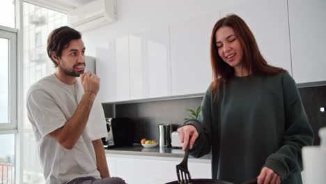 Una-Chica-Morena-Feliz-Con-Un-Suéter-Verde-Prepara-El-Desayuno-Mientras-Su-Novio,-Un-Hombre-Moreno-Con-Un-Escudo-Y-Una-Camiseta-Blanca,-Se-Apura-Los-Dientes-En-La-Cocina-De-Un-Apartamento-Moderno.